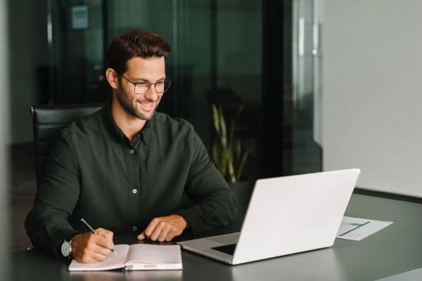 Smiling businessman in glasses taking notes while working on a laptop in a modern office.